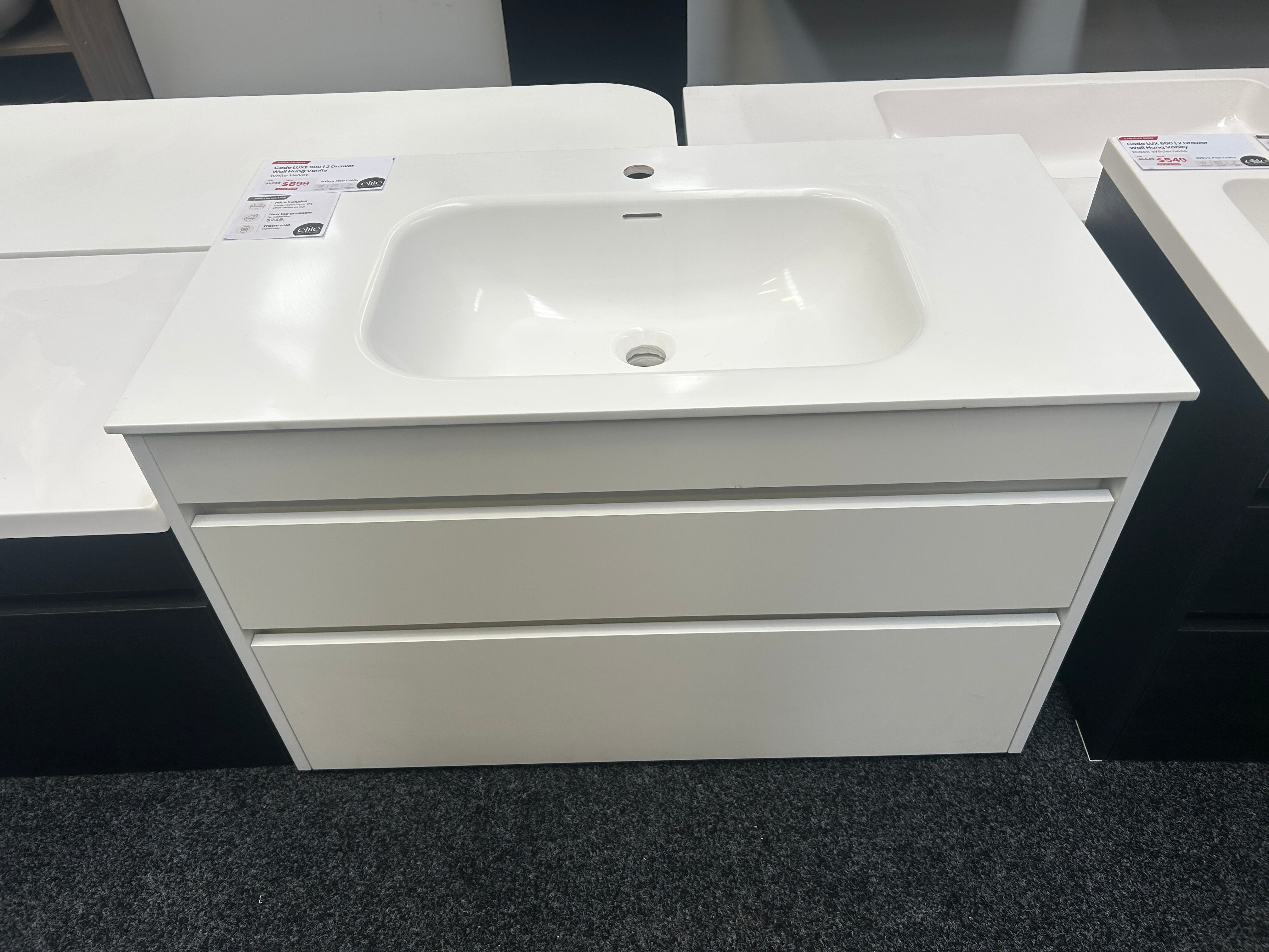A modern white bathroom vanity cabinet with a built-in rectangular sink, shown in a showroom setting with other sinks partially visible.