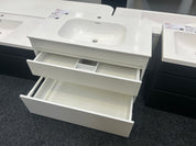 A white bathroom vanity cabinet with a sink basin and two open drawers, shown in a showroom setting with other similar units partially visible.