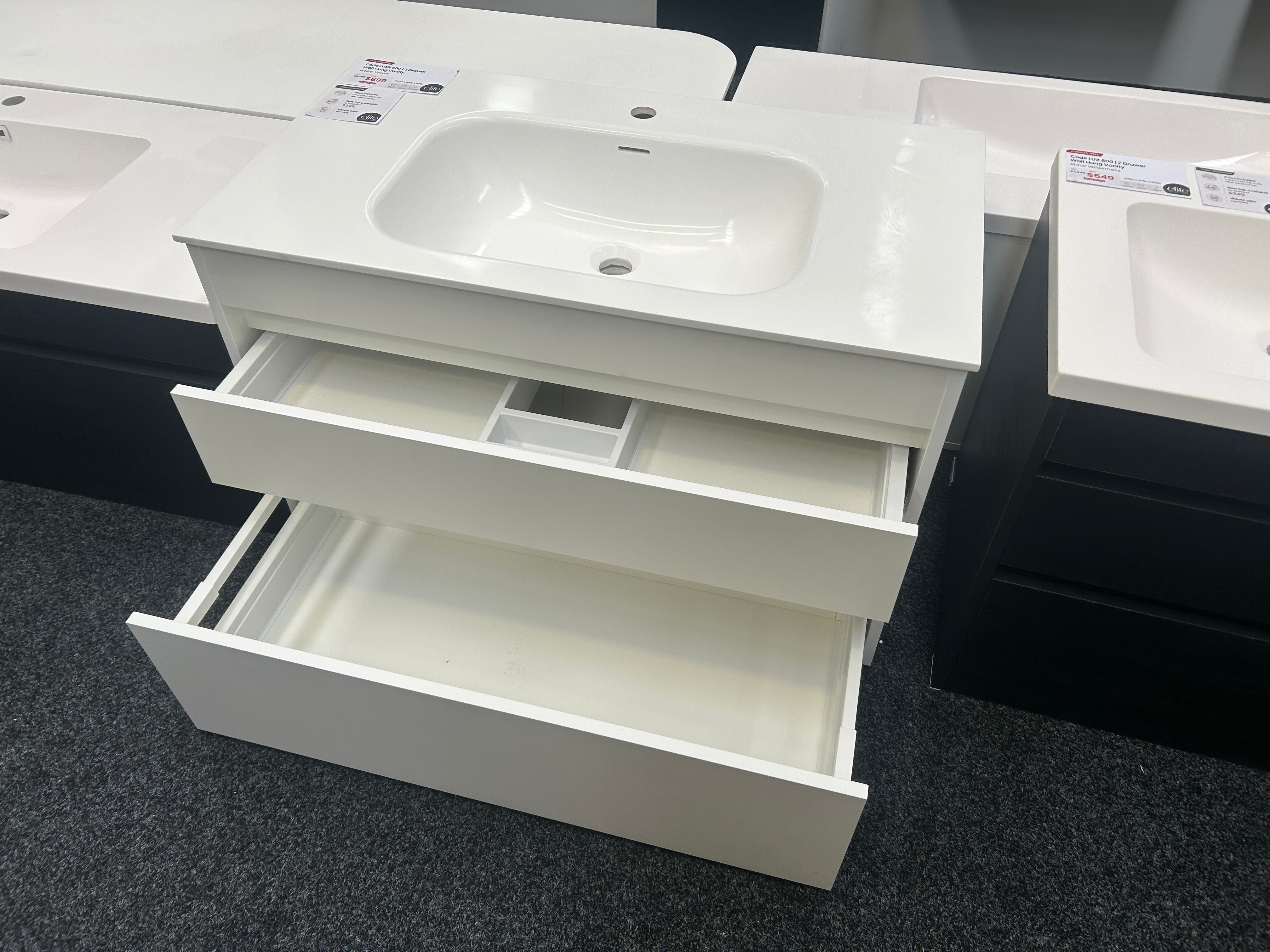A white bathroom vanity cabinet with a sink basin and two open drawers, shown in a showroom setting with other similar units partially visible.