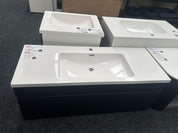 A bathroom vanity set with a white rectangular basin and countertop, paired with a dark charcoal/black cabinet, shown in a showroom with other similar units behind it.