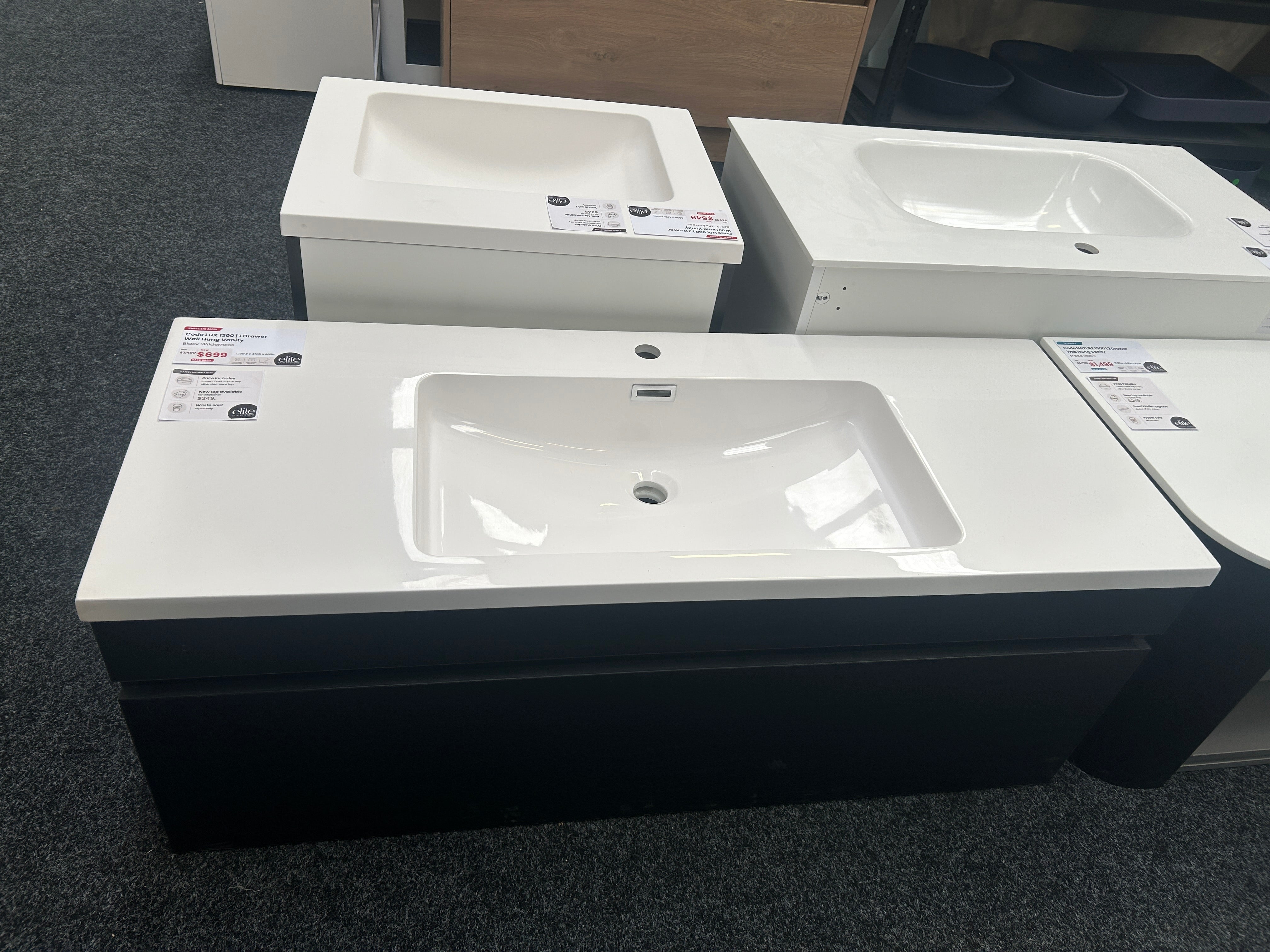 A bathroom vanity set with a white rectangular basin and countertop, paired with a dark charcoal/black cabinet, shown in a showroom with other similar units behind it.