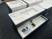 A modern bathroom vanity with a white rectangular sink countertop and dark charcoal cabinet, shown with the lower drawer pulled open on a dark carpeted showroom floor.