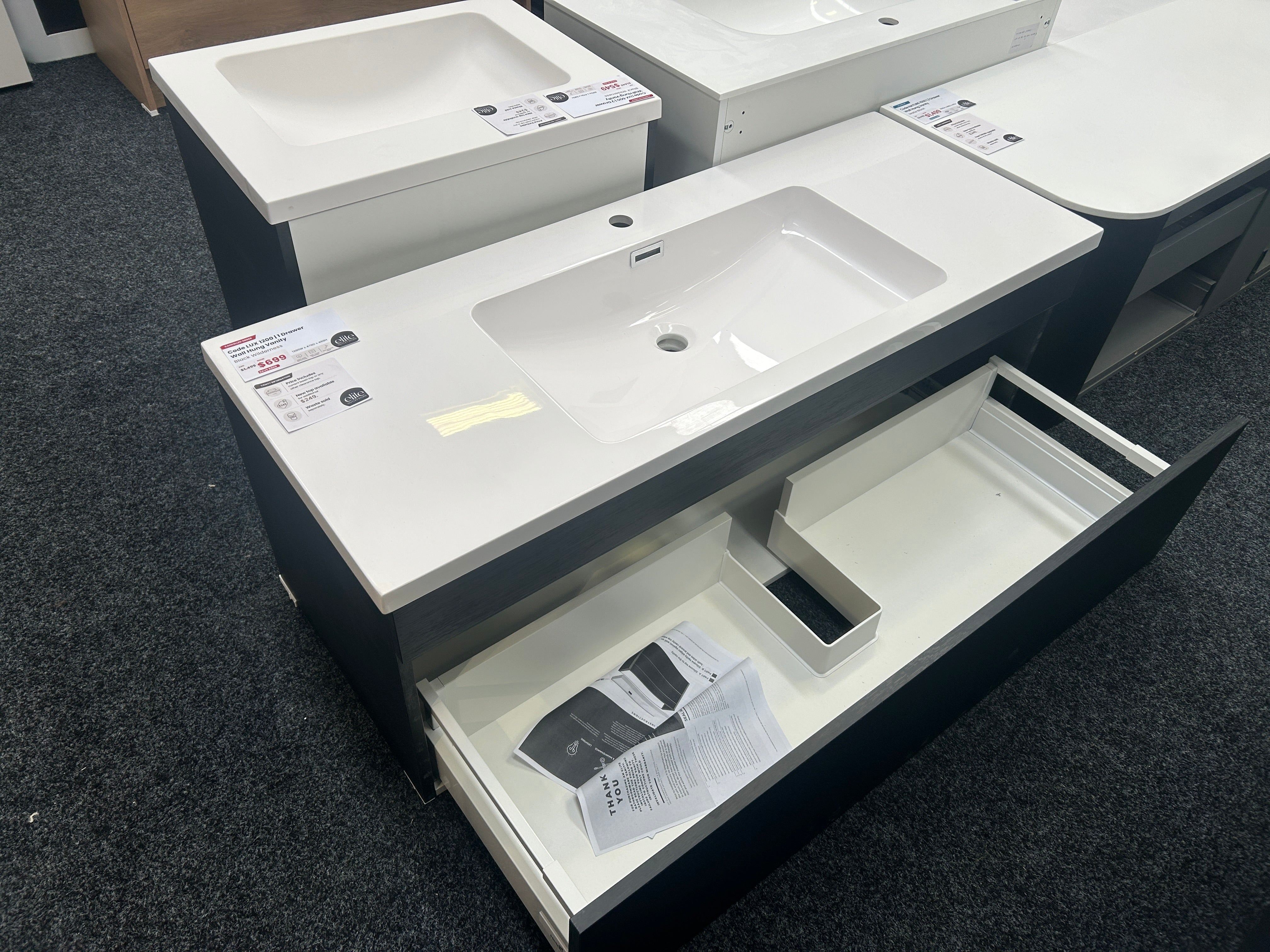 A modern bathroom vanity with a white rectangular sink countertop and dark charcoal cabinet, shown with the lower drawer pulled open on a dark carpeted showroom floor.