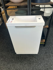 A white pedestal sink with a rectangular basin and a horizontal metallic handle, shown in a showroom or warehouse setting with shelving and other fixtures in the background.