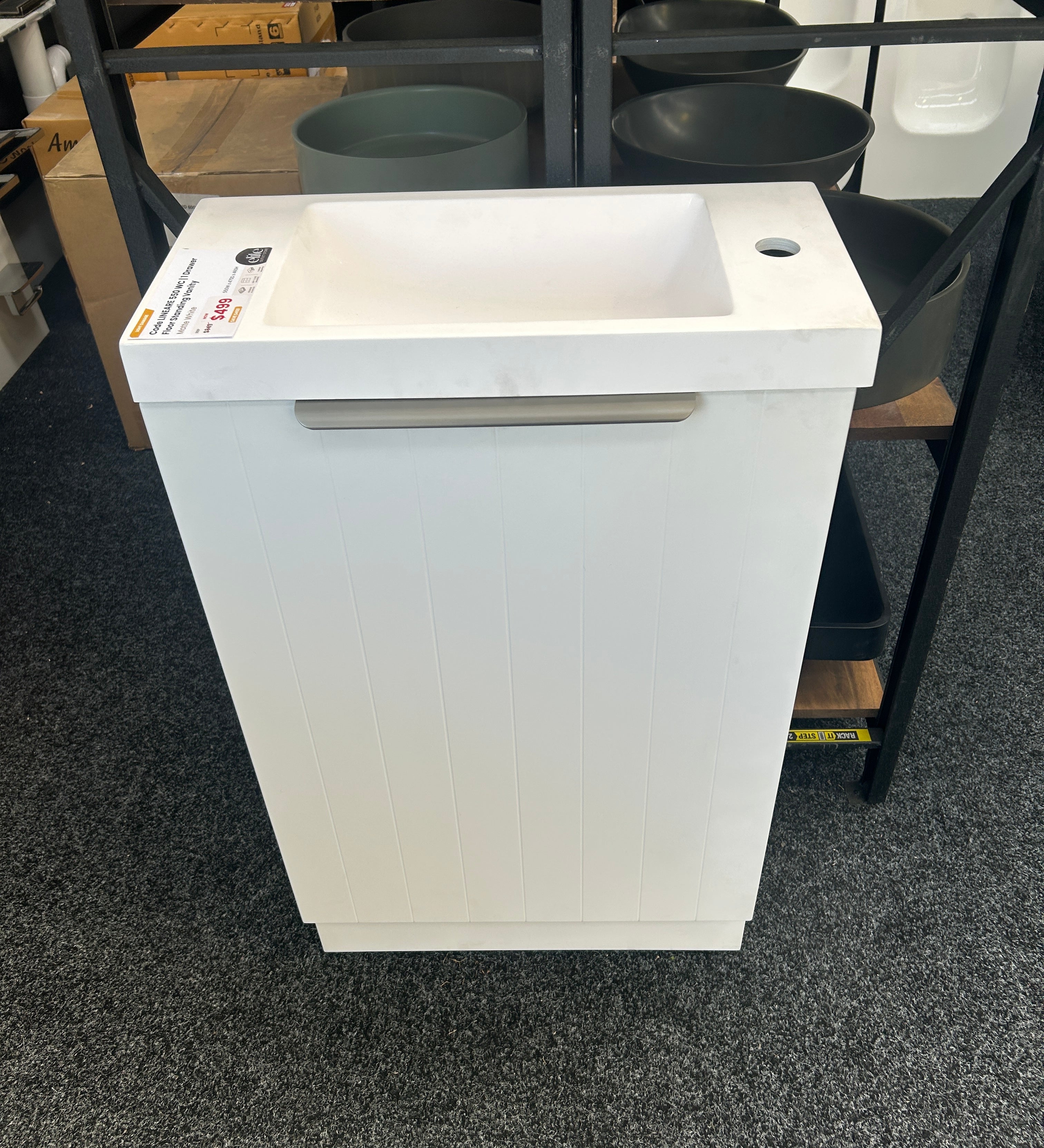 A white pedestal sink with a rectangular basin and a horizontal metallic handle, shown in a showroom or warehouse setting with shelving and other fixtures in the background.