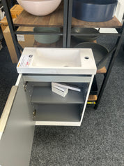 A white bathroom vanity cabinet with an integrated sink and open lower cabinet door, shown on a store floor with shelving in the background.