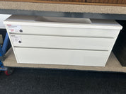 A white two-drawer undercounter cabinet with a built-in sink basin on top, shown in a warehouse or showroom setting with carpeted floors and a blue cart visible on the left.