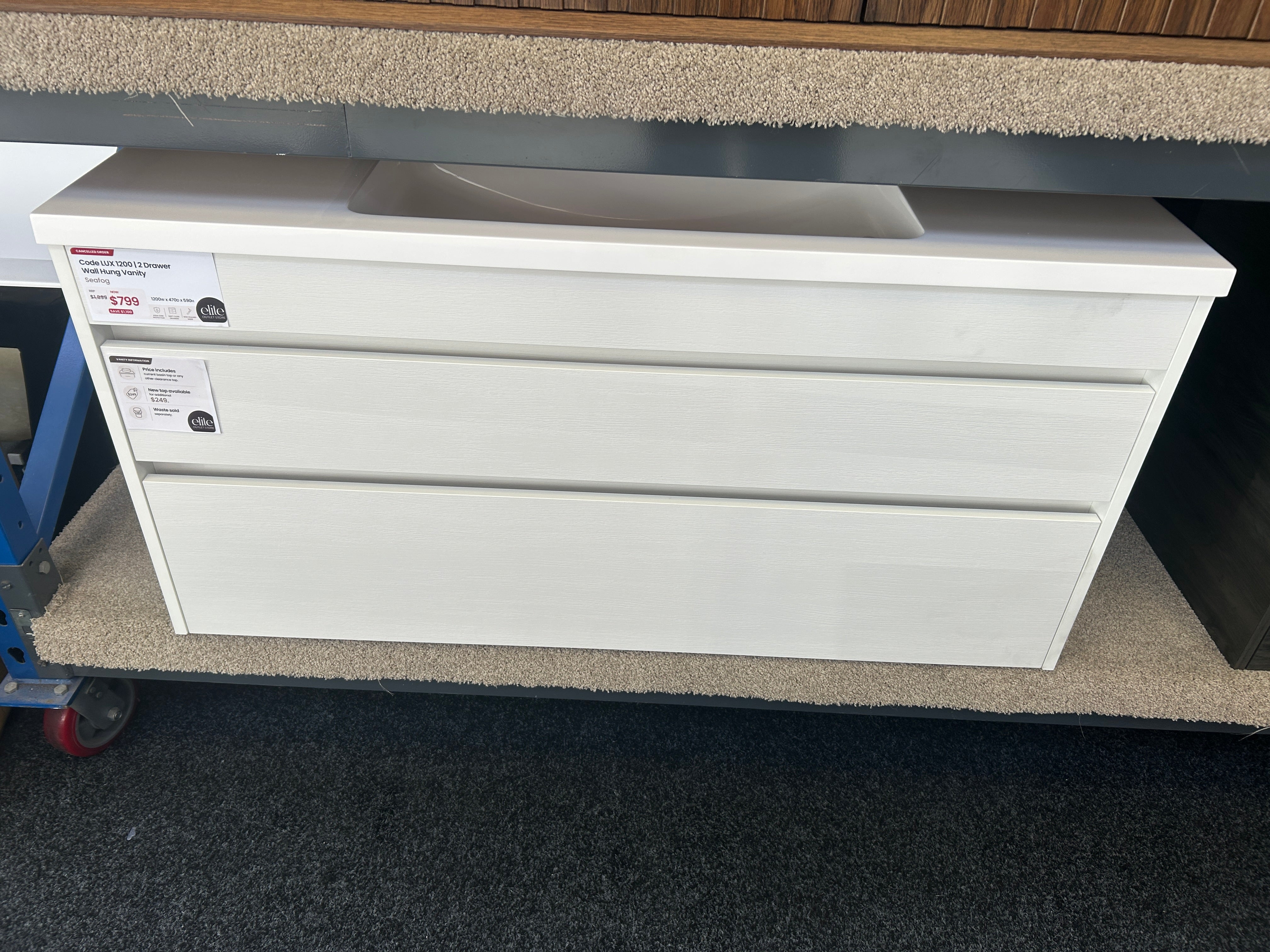 A white two-drawer undercounter cabinet with a built-in sink basin on top, shown in a warehouse or showroom setting with carpeted floors and a blue cart visible on the left.
