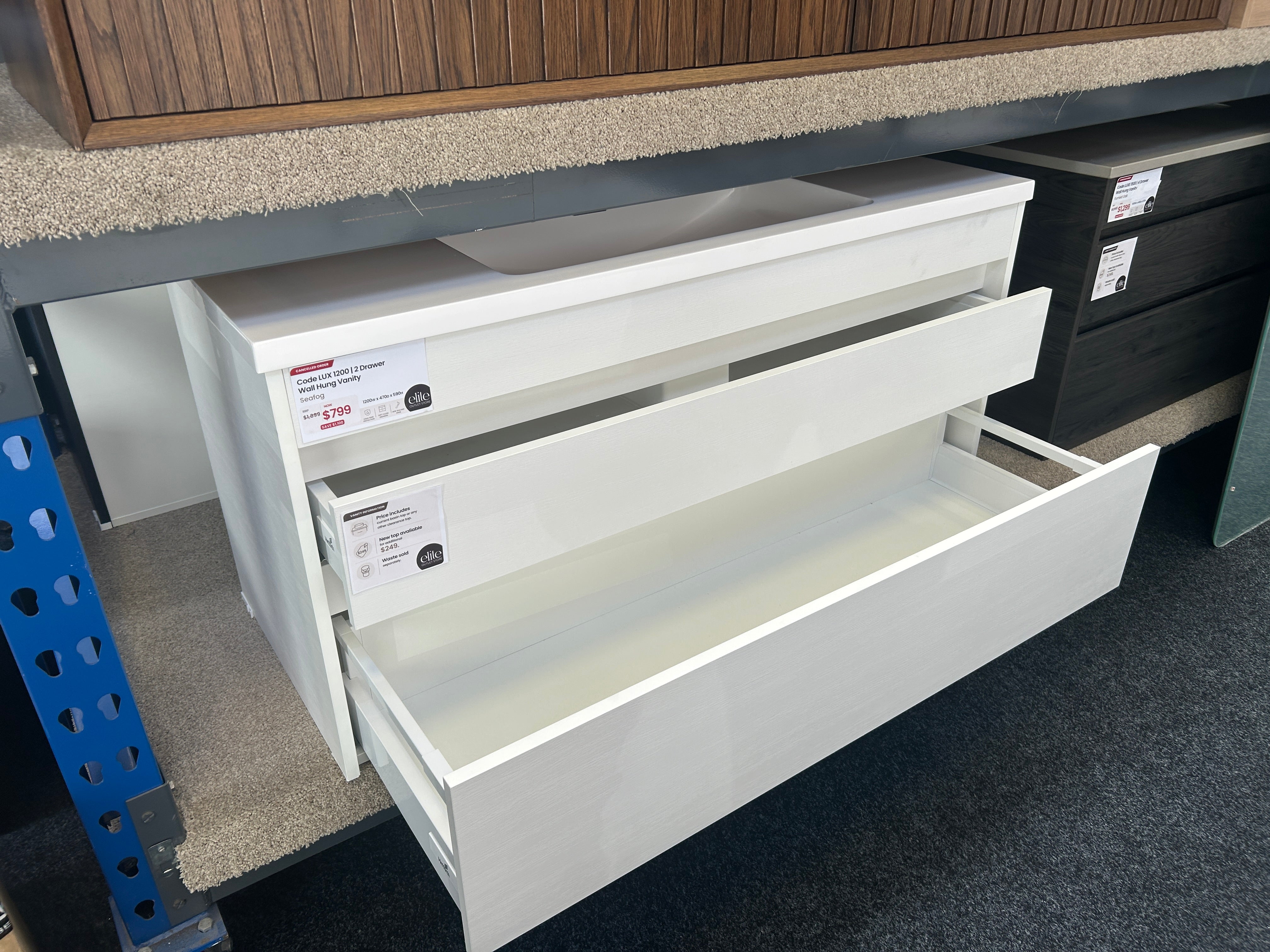 White drawer unit with two partially open drawers, photographed in a showroom or warehouse setting with carpeted floor and other furniture visible.