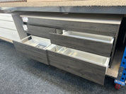 Gray wood-look drawer cabinet with multiple drawers, including two open drawers showing white interiors, photographed in a showroom or warehouse setting.