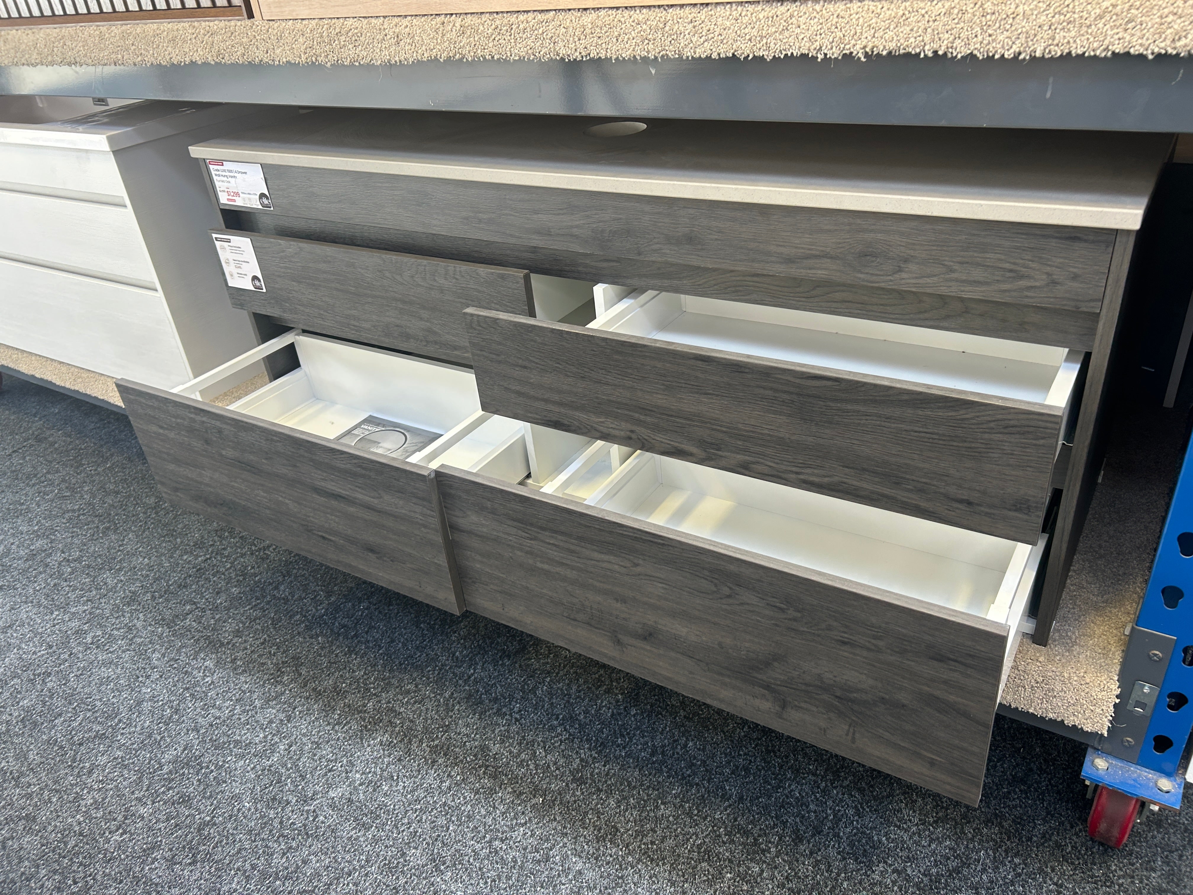 Gray wood-look drawer cabinet with multiple drawers, including two open drawers showing white interiors, photographed in a showroom or warehouse setting.