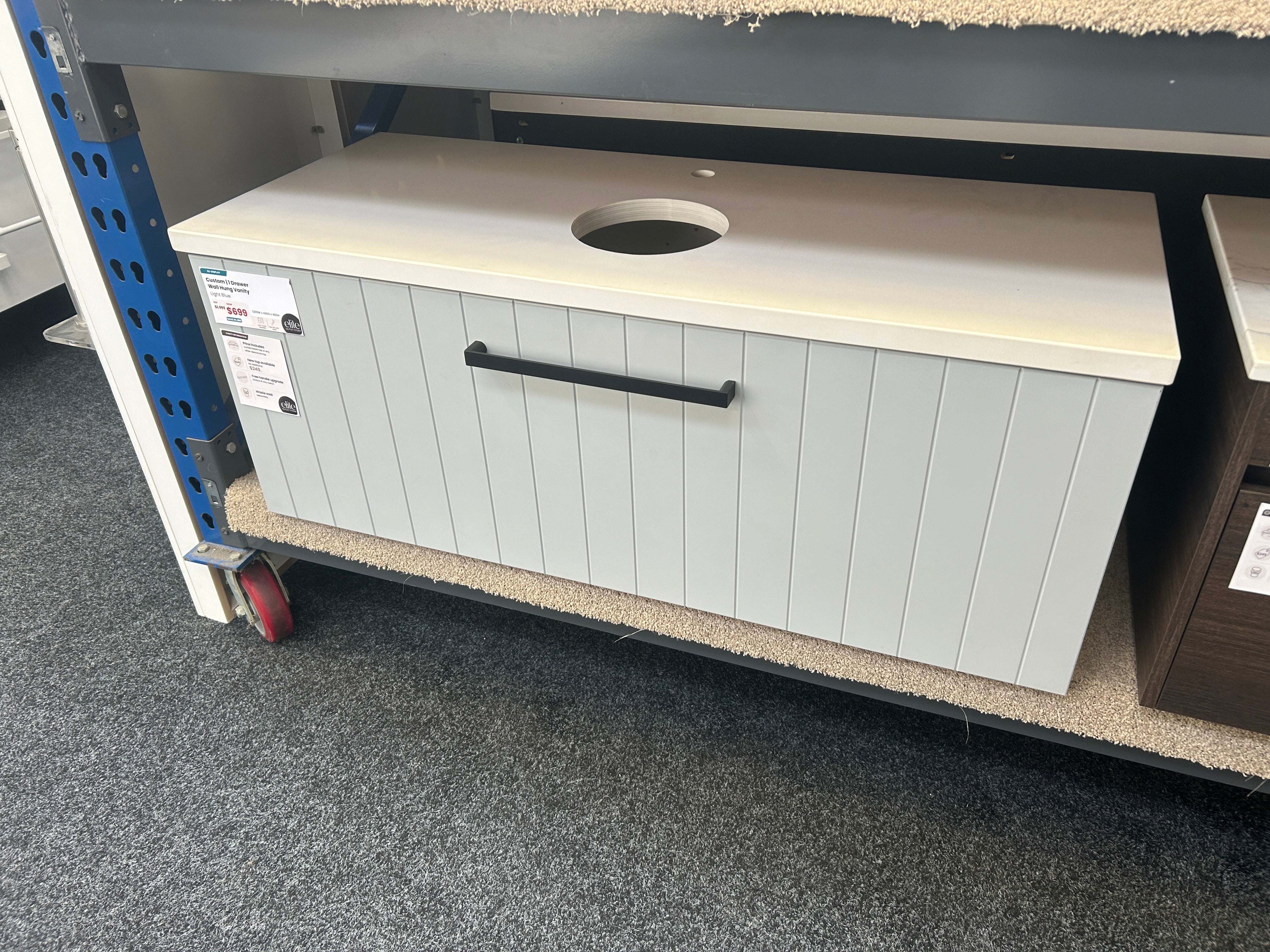 A light gray cabinet with a cream countertop and a black pull handle, photographed on a store shelf with carpeted floor and shelving visible.