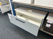 A white shiplap-style undercounter cabinet with a single black horizontal handle, shown open on a store display shelf with carpeted floors and other furniture pieces around it.