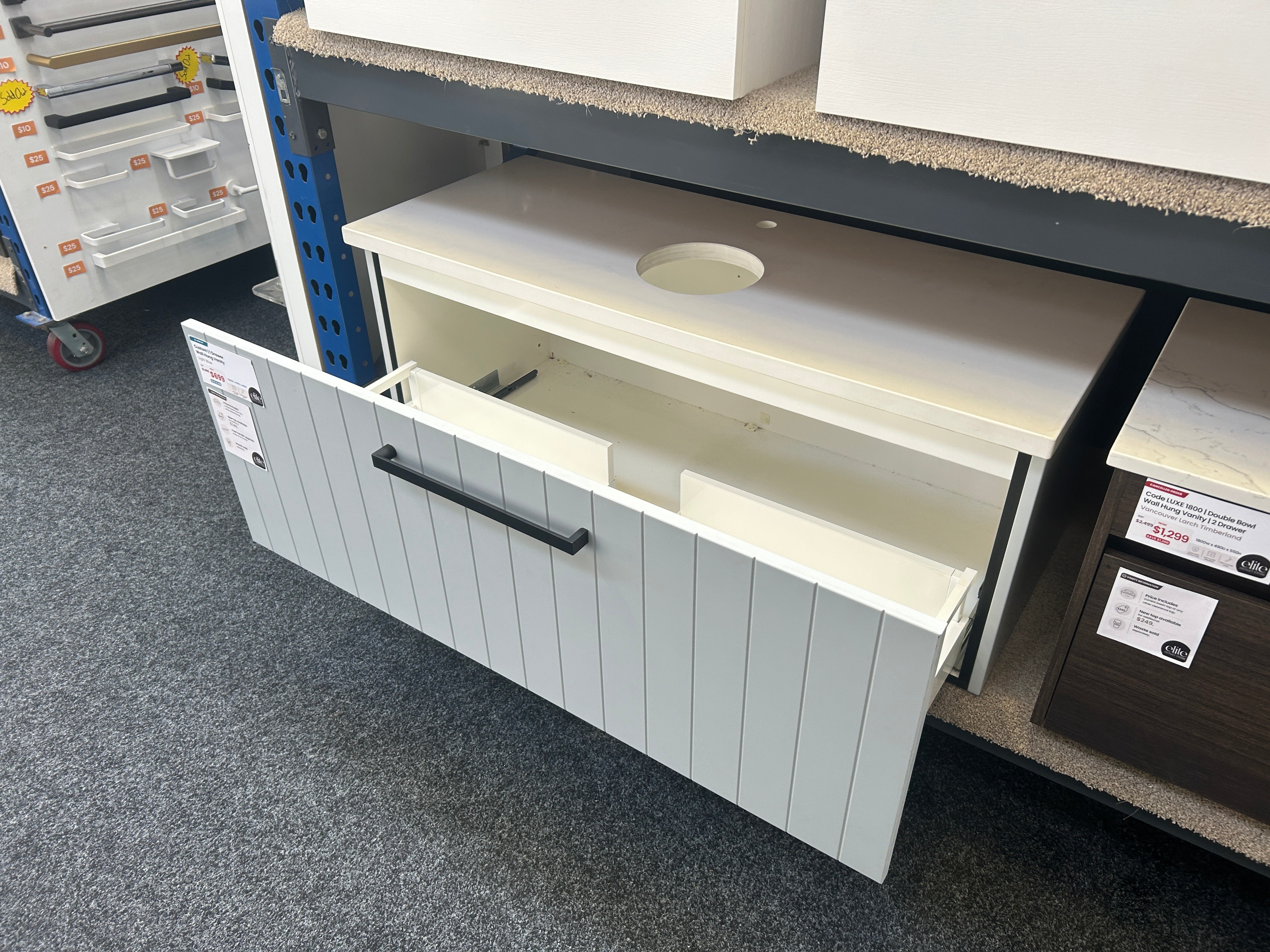 A white shiplap-style undercounter cabinet with a single black horizontal handle, shown open on a store display shelf with carpeted floors and other furniture pieces around it.