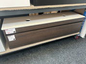 A long rectangular under-desk storage cabinet with a dark brown wood-grain finish and a light beige top surface, shown on a showroom floor under a desk.