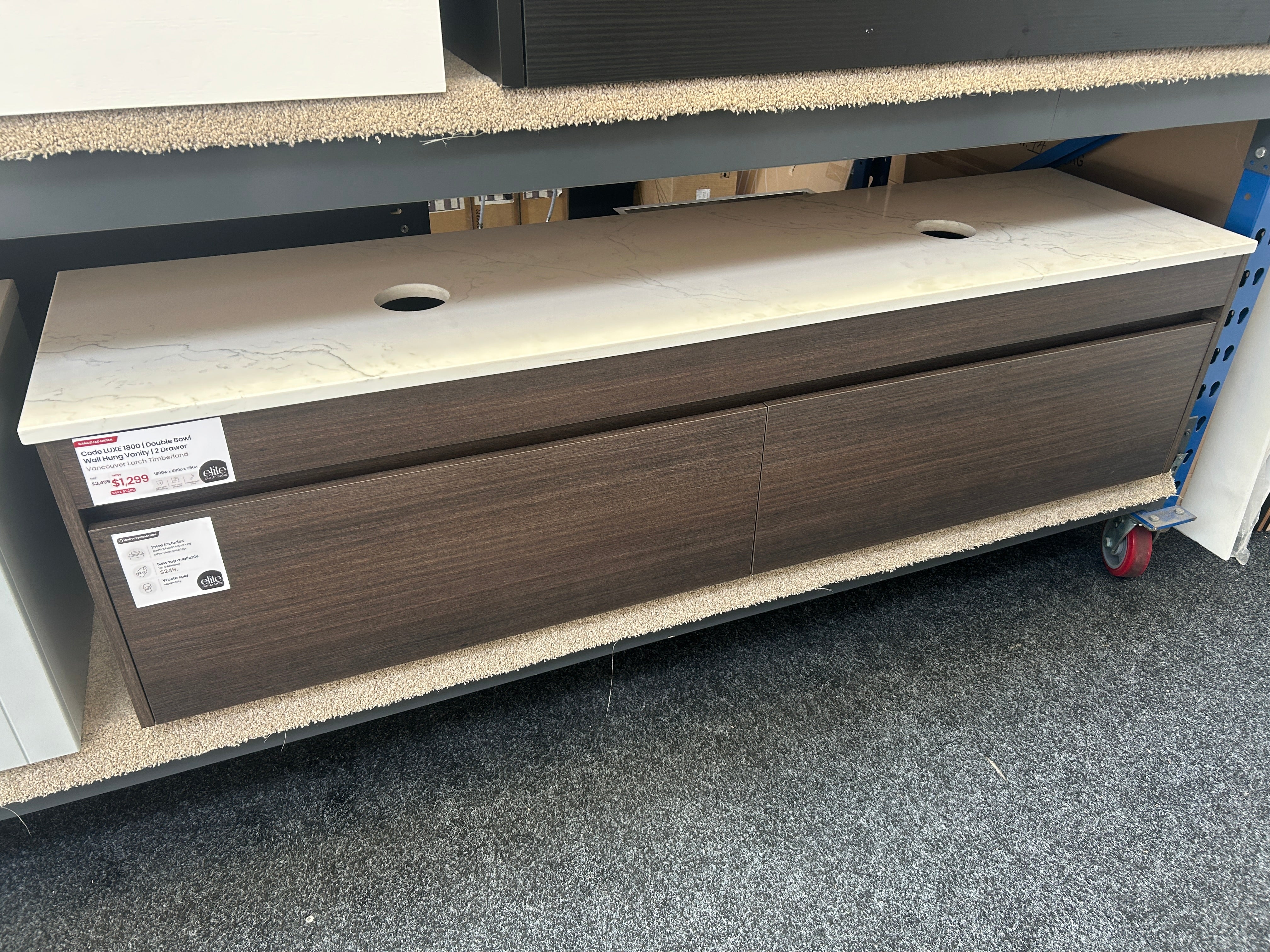 A long rectangular under-desk storage cabinet with a dark brown wood-grain finish and a light beige top surface, shown on a showroom floor under a desk.