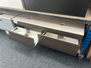 A modern vanity or storage cabinet with dark brown wood-grain fronts, a light stone-look top, and open drawers showing white interior organizers, photographed in a showroom or warehouse setting on gray carpet.