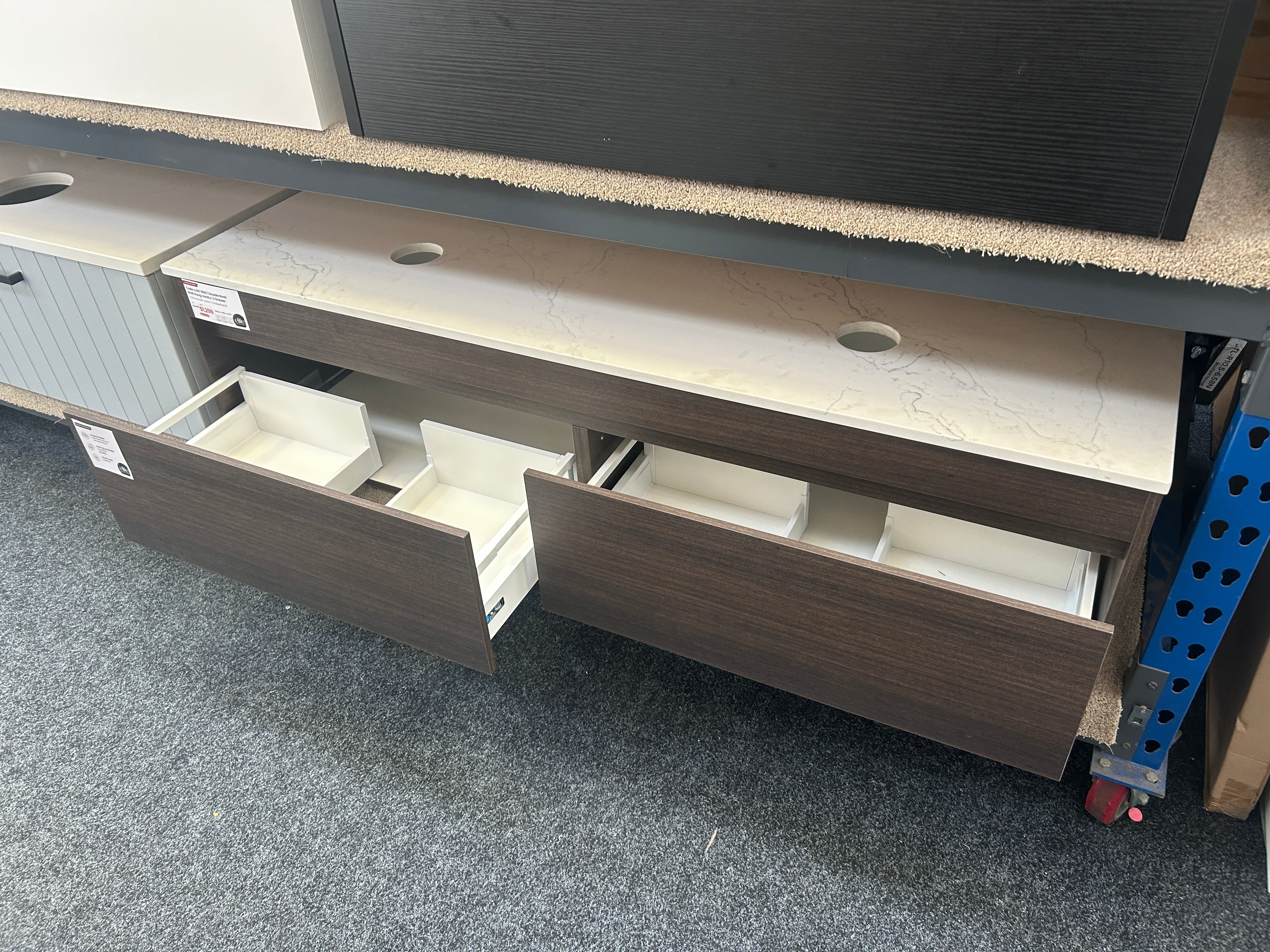 A modern vanity or storage cabinet with dark brown wood-grain fronts, a light stone-look top, and open drawers showing white interior organizers, photographed in a showroom or warehouse setting on gray carpet.