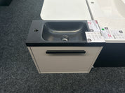 A modern wall-mounted bathroom vanity with a dark charcoal gray sink basin and a cream-colored cabinet door with a black handle, shown on a gray carpeted showroom floor.