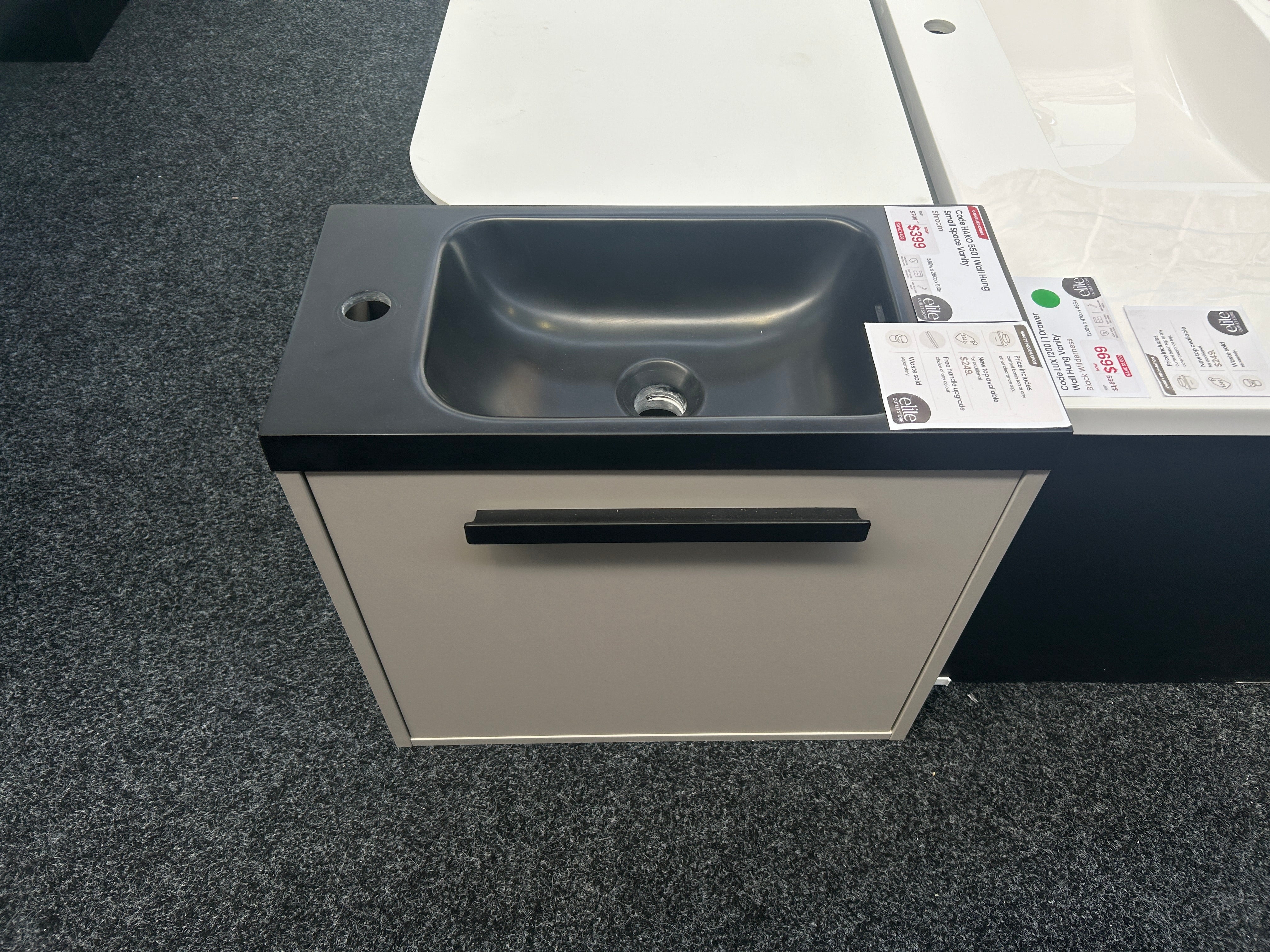 A modern wall-mounted bathroom vanity with a dark charcoal gray sink basin and a cream-colored cabinet door with a black handle, shown on a gray carpeted showroom floor.