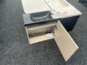 A compact bathroom vanity cabinet with an integrated sink, shown with one door open on a gray carpeted floor. The vanity has a cream-colored cabinet base, a dark charcoal sink basin, and a white countertop with a matching white sink inset.