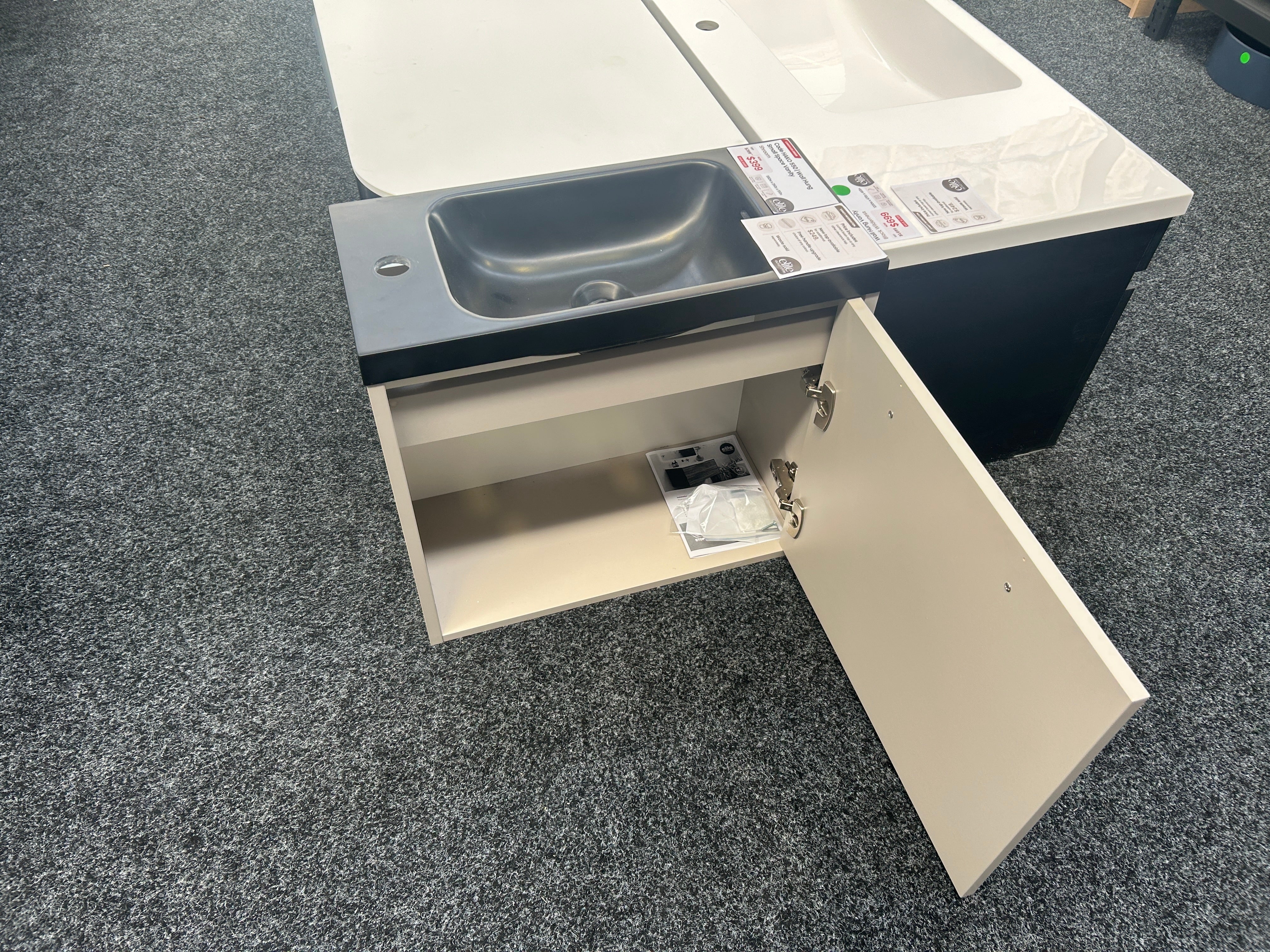 A compact bathroom vanity cabinet with an integrated sink, shown with one door open on a gray carpeted floor. The vanity has a cream-colored cabinet base, a dark charcoal sink basin, and a white countertop with a matching white sink inset.