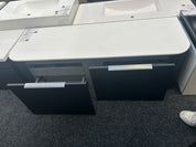 A modern bathroom vanity cabinet with a white countertop and dark charcoal/black drawer fronts, shown in a showroom with other sinks and a gray carpeted floor.