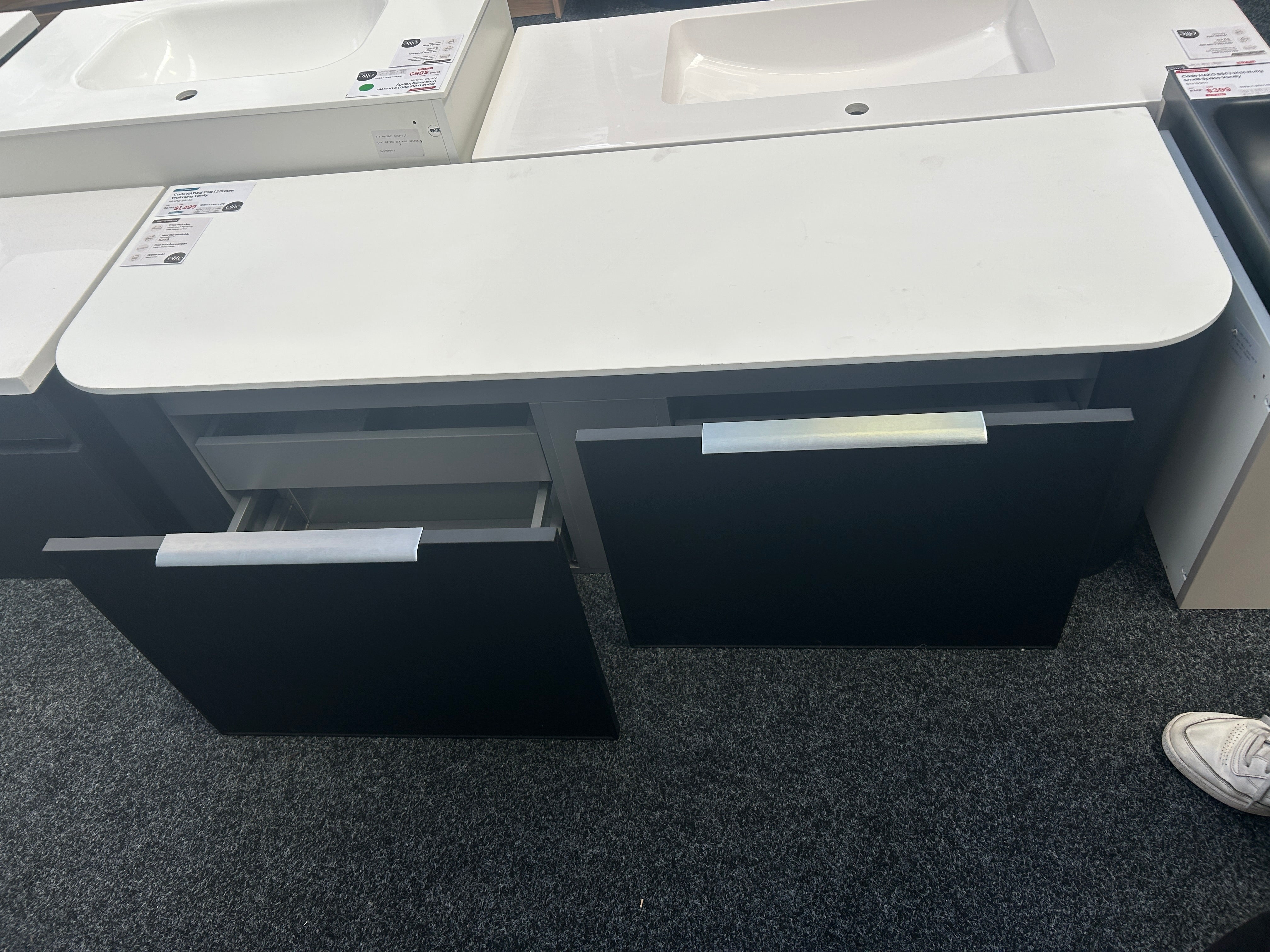 black drawer fronts, shown in a showroom with other sinks and a gray carpeted floor.
