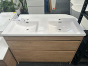 A double-sink bathroom vanity with a white countertop and integrated sinks, paired with a light oak wood cabinet, shown in a showroom setting.