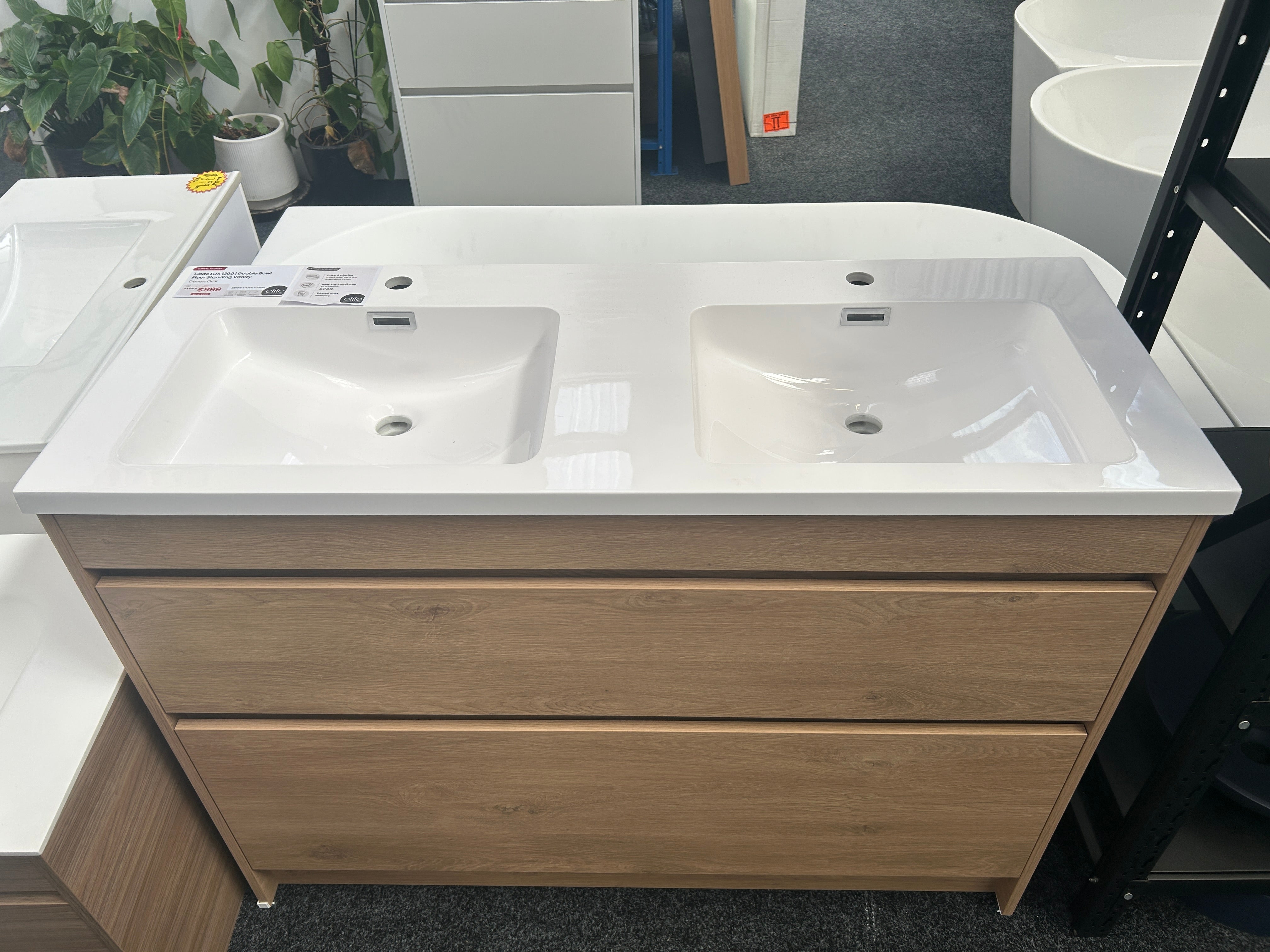 A double-sink bathroom vanity with a white countertop and integrated sinks, paired with a light oak wood cabinet, shown in a showroom setting.