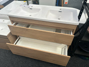 A double-sink bathroom vanity cabinet with a white ceramic countertop and integrated sinks, light wood grain doors and drawers, with two lower drawers pulled open, photographed in a showroom.