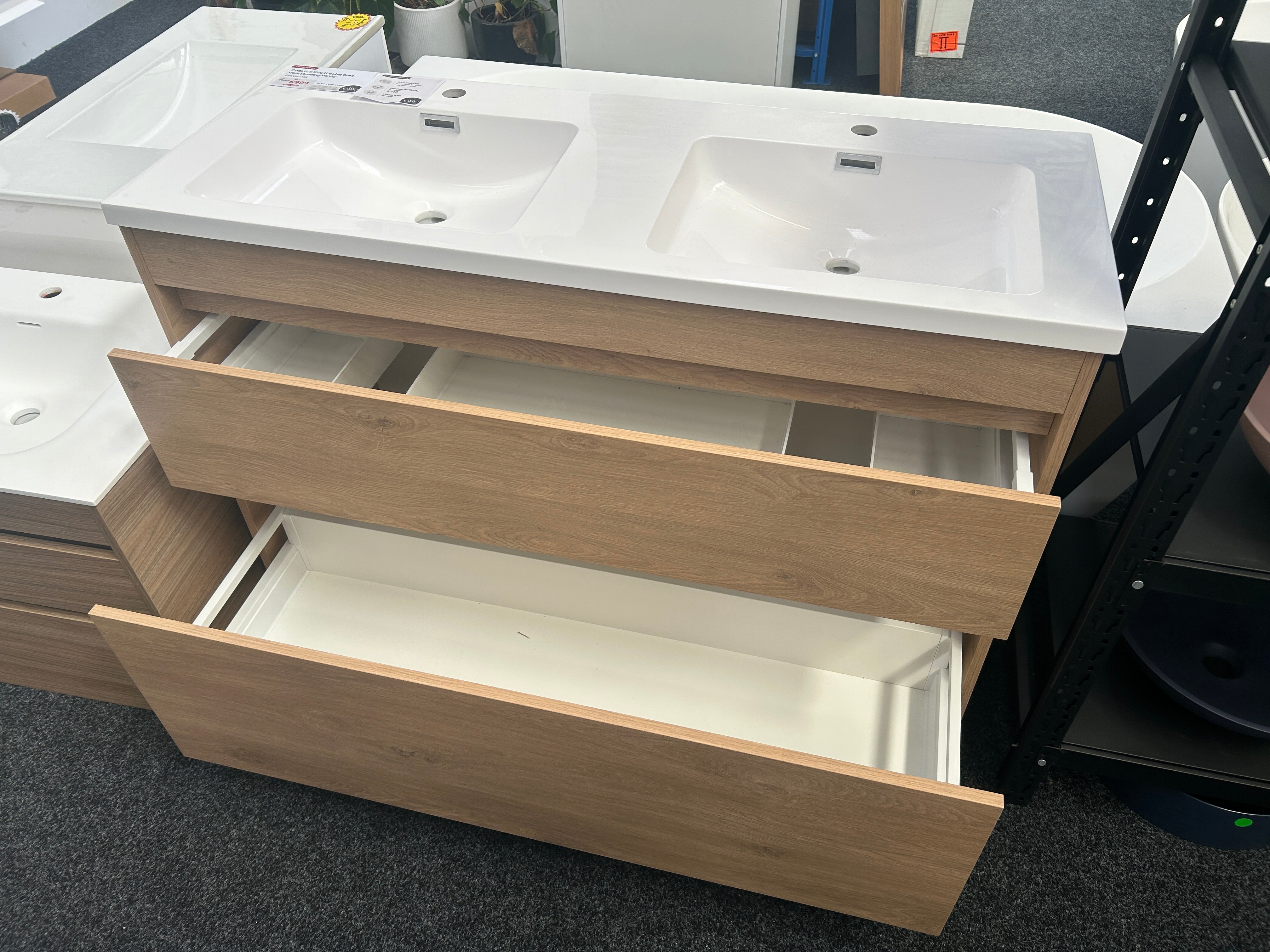 A double-sink bathroom vanity cabinet with a white ceramic countertop and integrated sinks, light wood grain doors and drawers, with two lower drawers pulled open, photographed in a showroom.