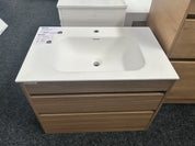 A modern bathroom vanity with a white integrated sink and a light brown wood-grain cabinet, shown on a dark gray carpeted showroom floor.