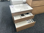 A modern bathroom vanity cabinet with a white integrated sink and two open wood-grain drawers, shown on a dark gray carpeted showroom floor.