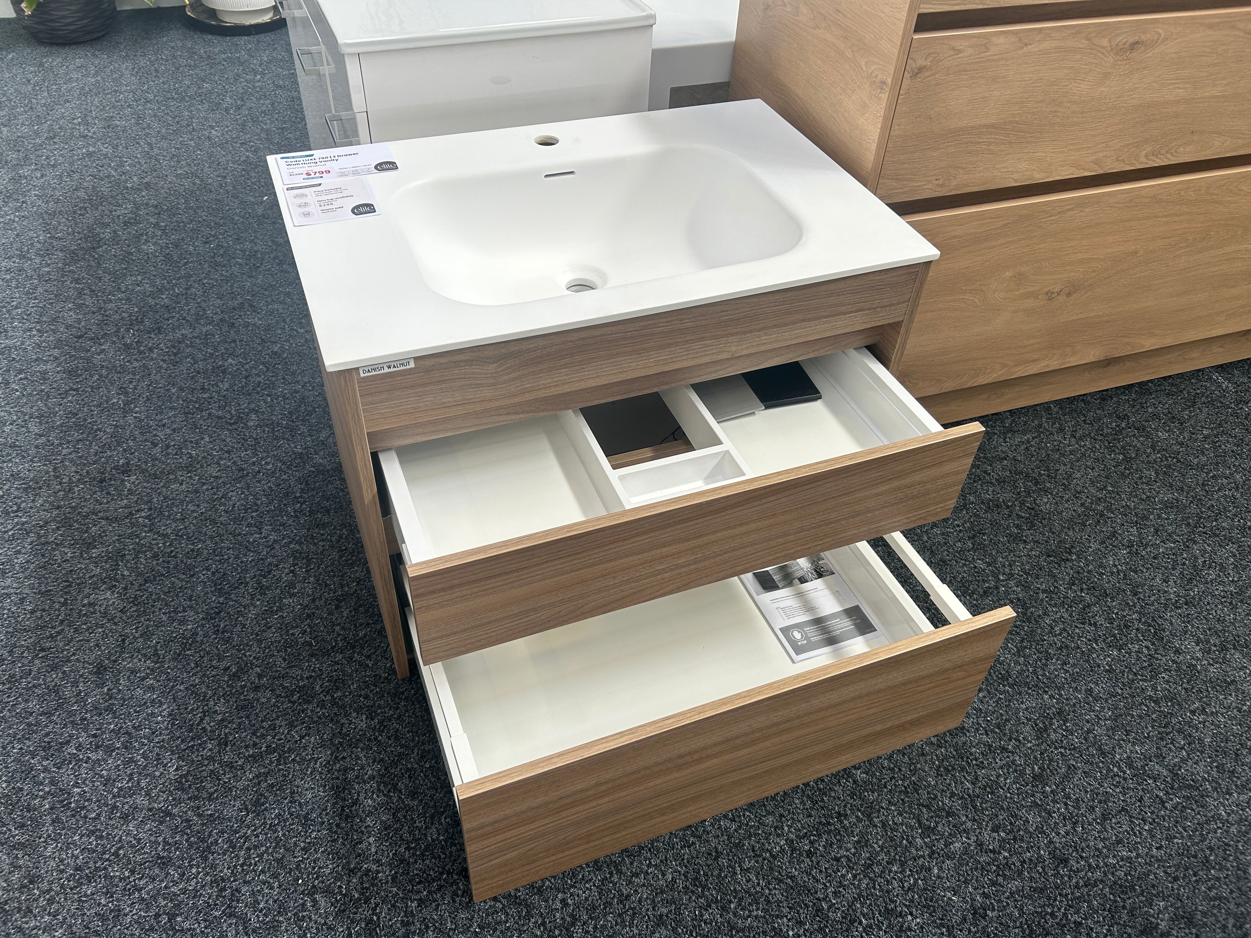 A modern bathroom vanity cabinet with a white integrated sink and two open wood-grain drawers, shown on a dark gray carpeted showroom floor.
